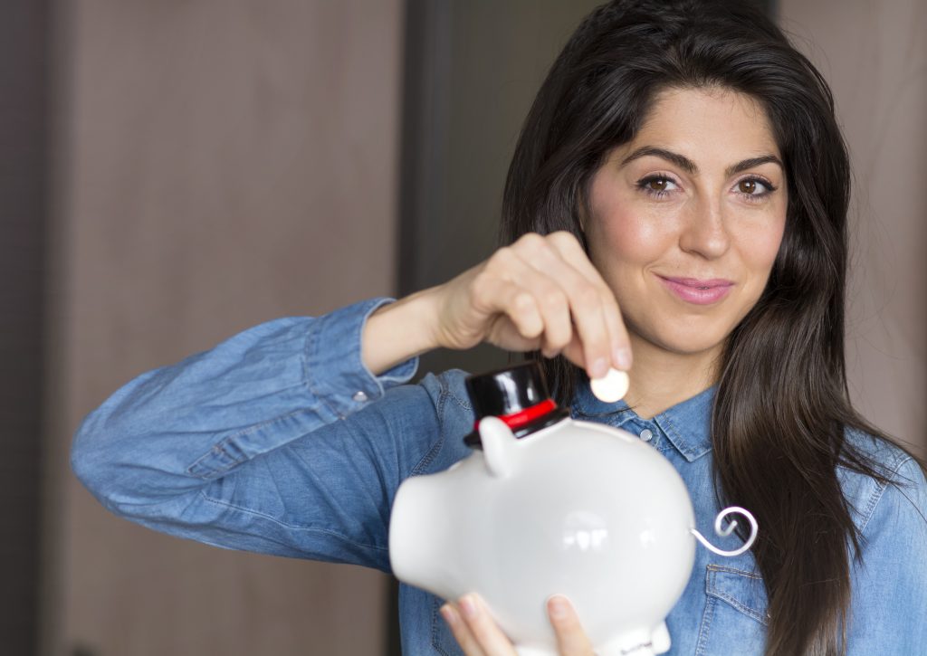 A young woman with long dark hair and a denim shirt is smiling while holding a white piggy bank with a small black hat and a curled tail. She is inserting a coin into the piggy bank, symbolizing saving money. Her facial expression conveys confidence and satisfaction. The background is blurred, focusing attention on the woman and the piggy bank.