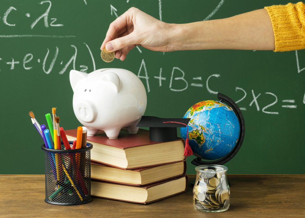 A person wearing a yellow sweater is placing a coin into a white piggy bank, which is positioned on top of a stack of three books. Next to the piggy bank, there is a small globe wearing a black graduation cap with a red tassel, symbolizing education and financial planning. A jar filled with coins sits on the right, while a black mesh pencil holder filled with colorful pens and brushes is on the left. The background features a green chalkboard with mathematical equations written in white chalk, reinforcing the theme of saving money for education.