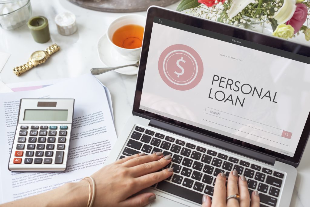 Woman using a laptop with a 'Personal Loan' webpage open, surrounded by financial documents, a calculator, a cup of tea, and flowers on a desk