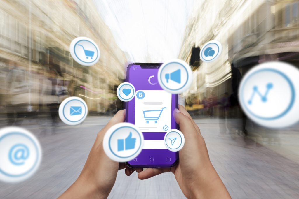 Person holding a smartphone with a shopping cart icon on the screen, surrounded by floating social media and e-commerce icons in a busy urban street background.
