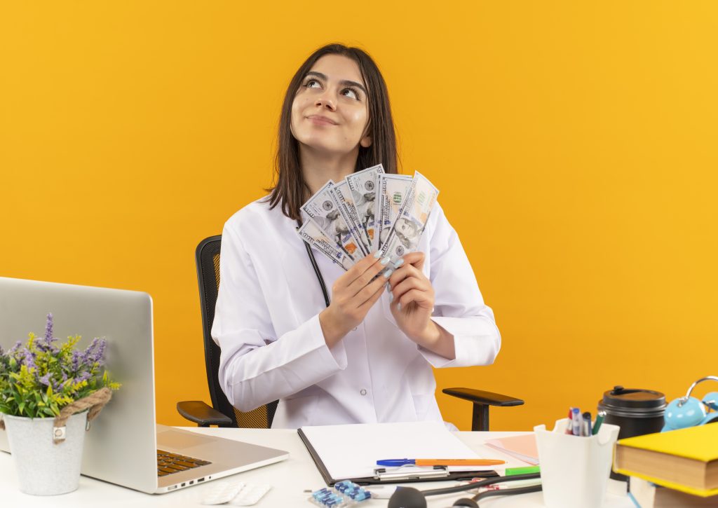 Young female doctor in a white coat holding a fan of U.S. dollar bills with a dreamy expression, sitting at a desk with a laptop, office supplies, and medical items against a bright orange background.