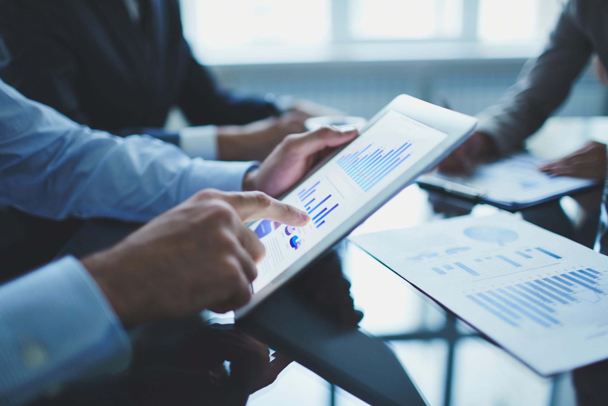 A close-up image showing a businessman using a digital tablet to analyze financial data, including bar and pie charts. The person is focused on reviewing important metrics and performance indicators on the screen, while others in the background are also engaged in a business discussion. This scene exemplifies the modern approach to managing and evaluating passive income opportunities through digital tools and data analysis.