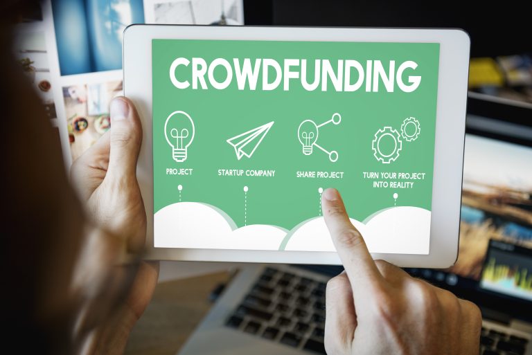 Close-up of a person using a tablet displaying a green screen with the word "Crowdfunding" and icons representing project creation, startup company, sharing a project, and turning ideas into reality.