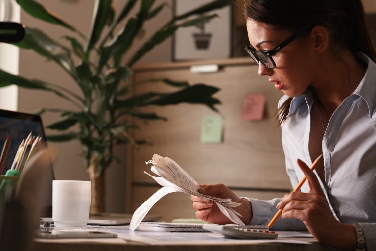 Focused woman wearing glasses sits at a desk analyzing receipts and using a calculator, representing financial planning, budgeting, or managing personal and business expenses in a home office setting.