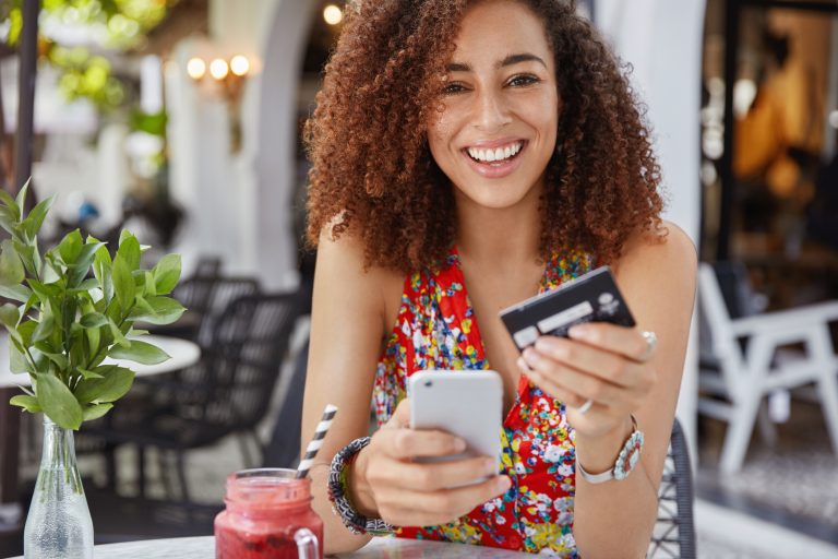 Smiling young woman sitting outdoors at a café, holding a smartphone and a credit card, symbolizing the convenience and mobility of online shopping with credit cards.