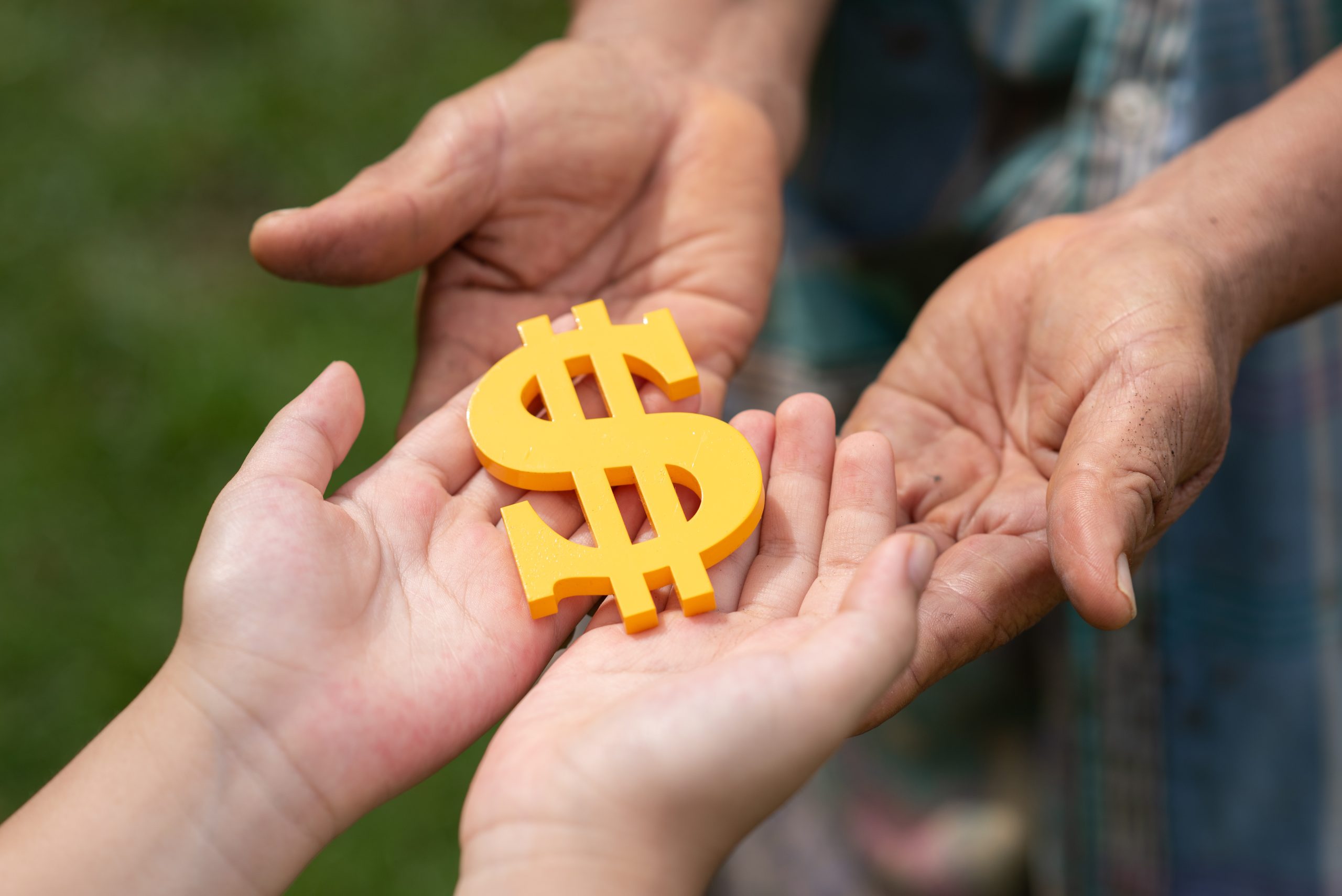 Close-up of multiple hands from different generations holding a yellow dollar sign, symbolizing financial collaboration and the concept of the sharing economy across age groups.