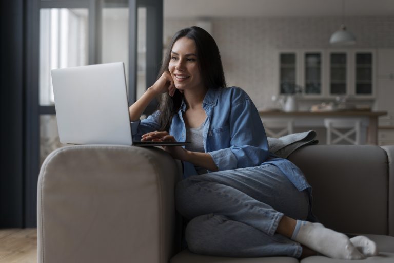 woman is sitting comfortably on a couch in a cozy living room, working on her laptop. She is smiling while looking at the screen, dressed casually in a denim shirt and jeans, with her legs crossed. The setting reflects a relaxed home office environment, emphasizing the flexibility of remote work and the comfort of working from home.