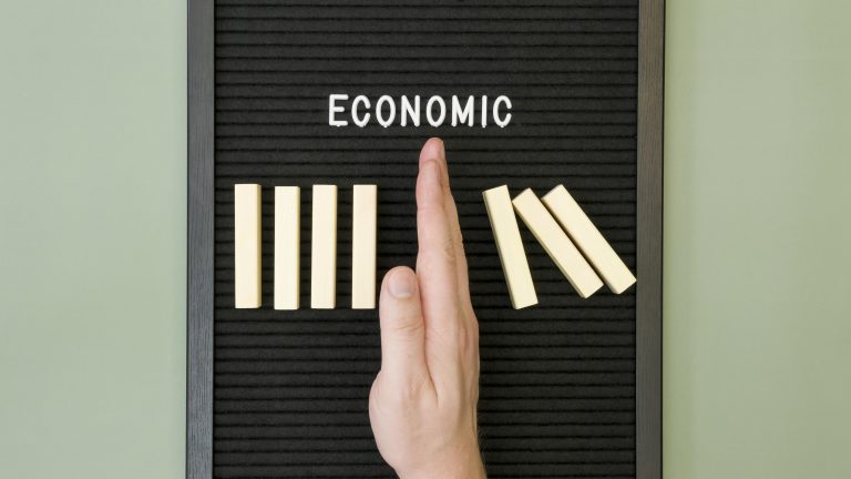 A hand stopping falling dominoes next to the word "ECONOMIC" on a black letter board, symbolizing efforts to prevent economic inequality from worsening.
