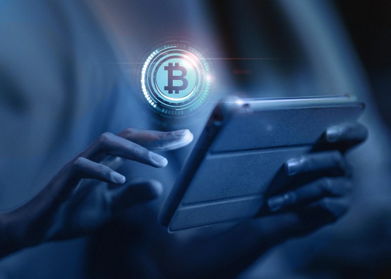 A person interacts with a digital tablet, with a glowing Bitcoin symbol holographically projected above the screen, representing the concept of digital currencies and cryptocurrency technology.