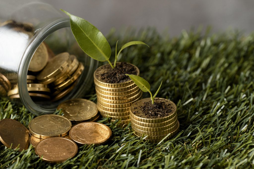 A glass jar spills golden coins onto grass, with two neat stacks of coins topped with green seedlings, symbolizing the growth potential of sustainable investments through environmentally conscious financial practices.