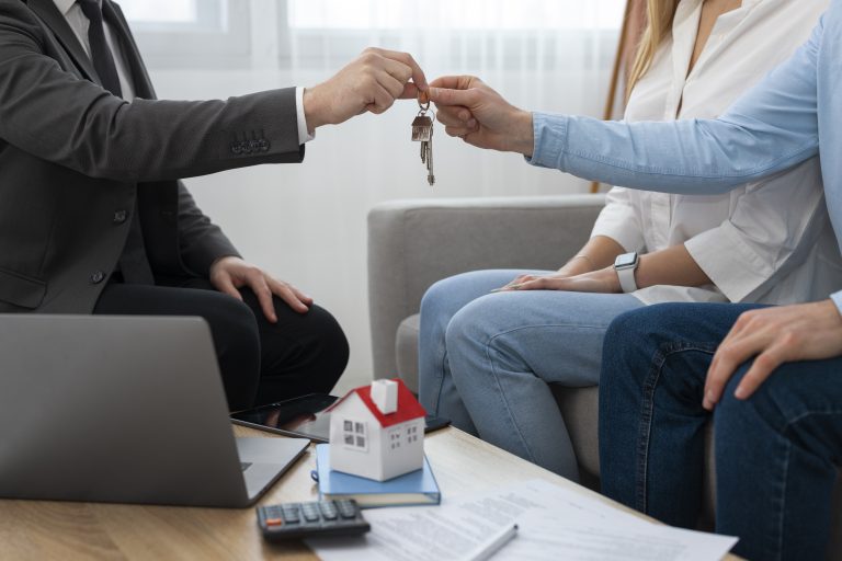 A real estate agent hands over house keys to a couple during a property transaction meeting, symbolizing a successful deal in the estate market. A miniature house model, laptop, documents, and calculator are visible on the table.