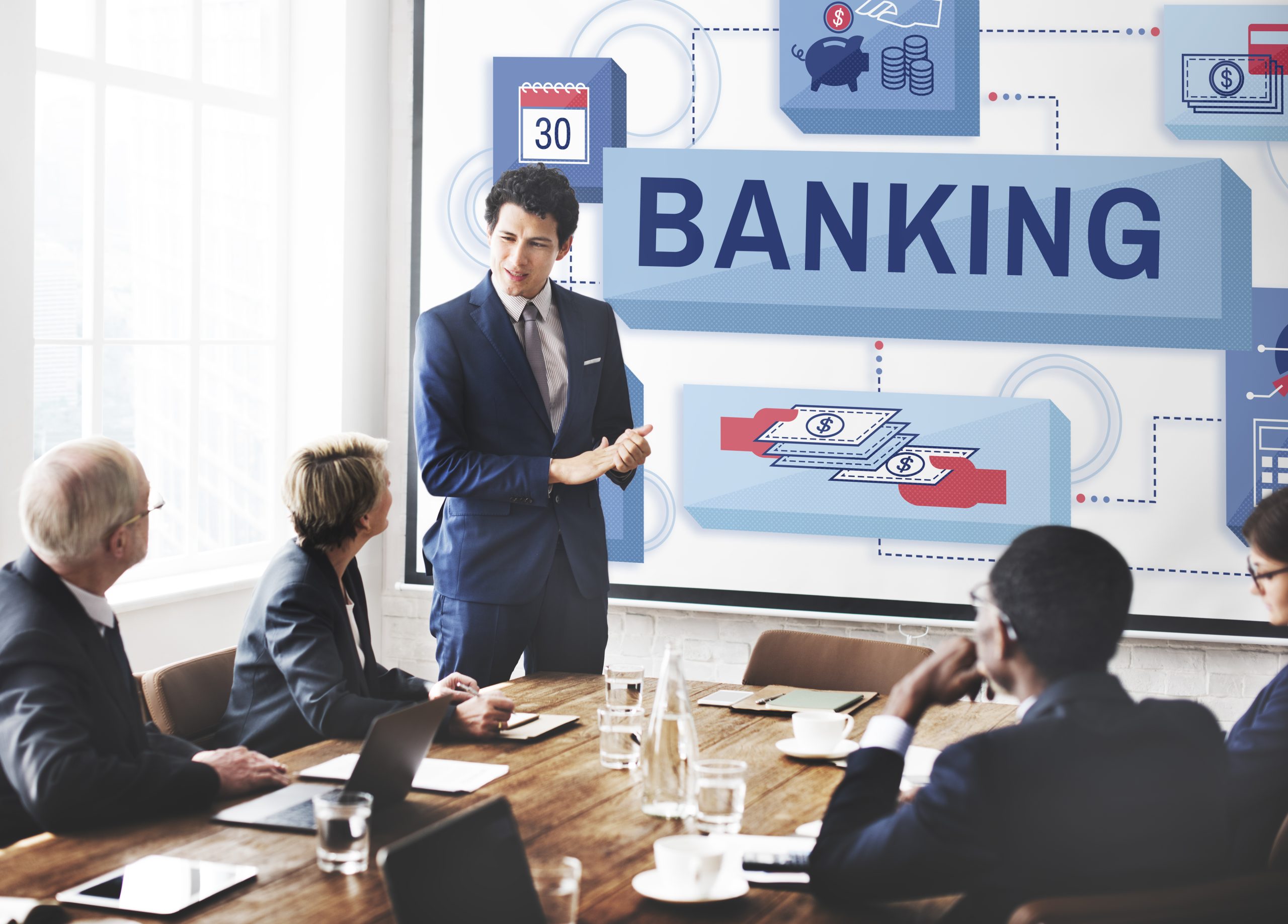 A business meeting in a modern office where a man in a blue suit is presenting a strategy about the banking sector to a diverse group of professionals, with a large digital screen displaying banking-related graphics and the word "BANKING" prominently featured.