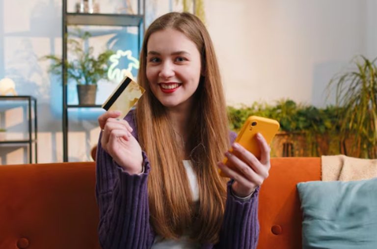 Smiling young woman sitting on a sofa holding a credit card and a smartphone, representing modern banking and digital payment solutions in the concept of Cards for Generation Z.