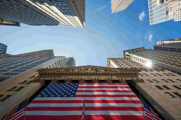 Classic façade on Wall Street with the United States flag, surrounded by modern skyscrapers under a blue sky, symbolizing the influence of the US Treasury and the American financial market.