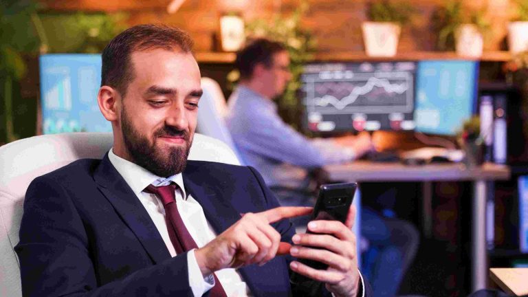 Businessman in a suit smiling while checking his smartphone, using social media for investment insights, with stock market charts displayed in the background.