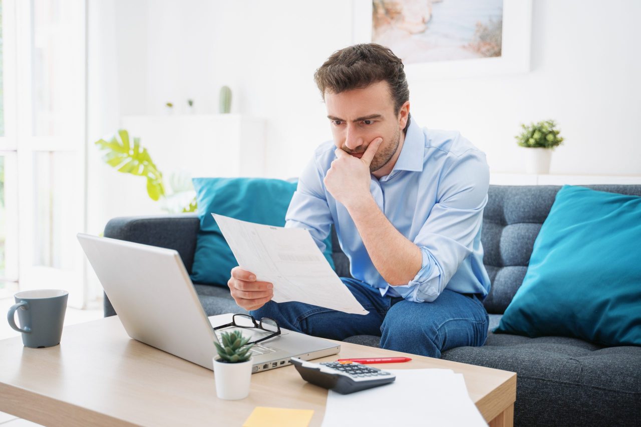 how to pay off debt in 2025: Man sitting on a couch at home looking worried while reviewing bills and financial documents, with a laptop and calculator on the table, representing financial stress and debt management.