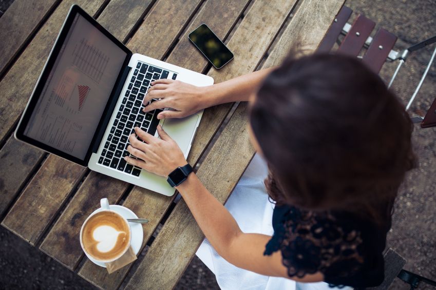 A woman works on a laptop displaying financial charts at an outdoor café table with coffee beside her, illustrating flexibility and convenience offered by the best online banks for beginners.