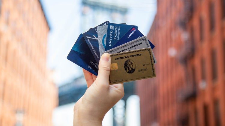 Hand holding several premium credit cards, including American Express and United Explorer, against an urban background — representing the best travel credit cards for rewards, miles, and travel benefits.
