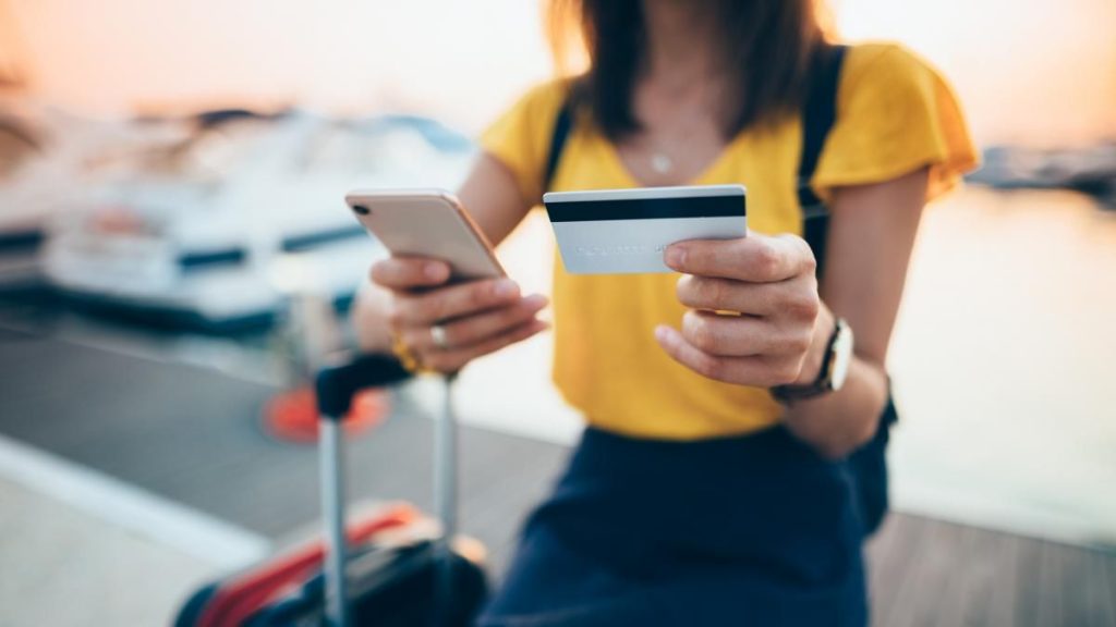 Traveler sitting with luggage at a marina, holding a smartphone and a credit card — illustrating how the best travel credit cards make it easy to earn cashback, miles, and rewards while on the go.