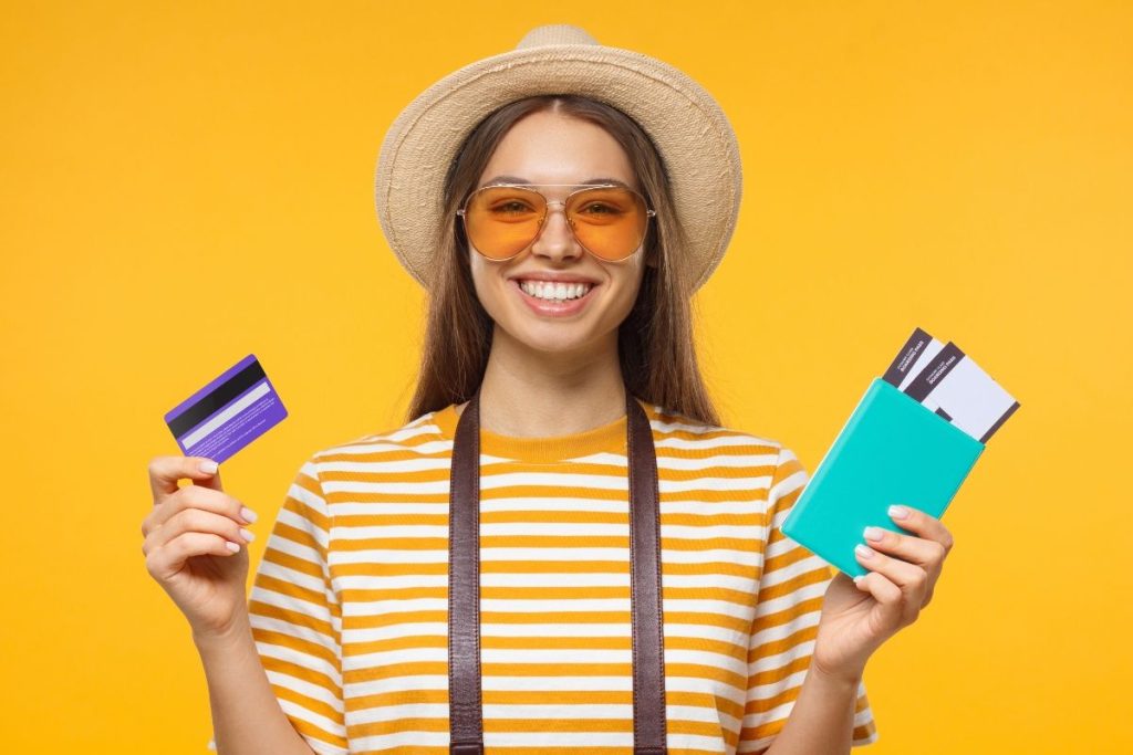 Smiling woman wearing a hat and sunglasses holding a credit card and passport with boarding passes — representing travelers who use the best travel credit cards to earn miles and exclusive perks.