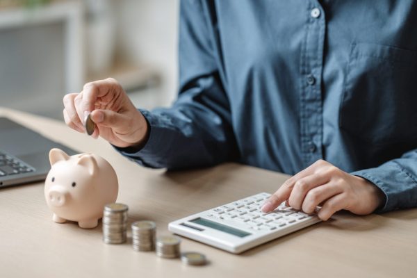 Person placing a coin into a piggy bank while using a calculator at a desk, illustrating practical budgeting habits and how to save money fast.
