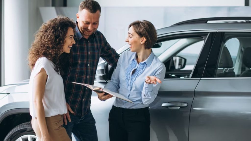 Car dealership consultant explaining auto loan vs leasing differences to a couple standing next to a new vehicle while reviewing financing options.