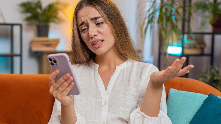 Woman sitting on a couch looking worried at her phone, illustrating the question “why was my loan application denied” after receiving a loan rejection.
