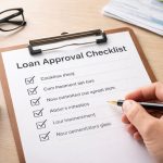 Hand holding a pen while checking items on a loan approval checklist on a clipboard, with financial documents, glasses, and a smartphone on a desk